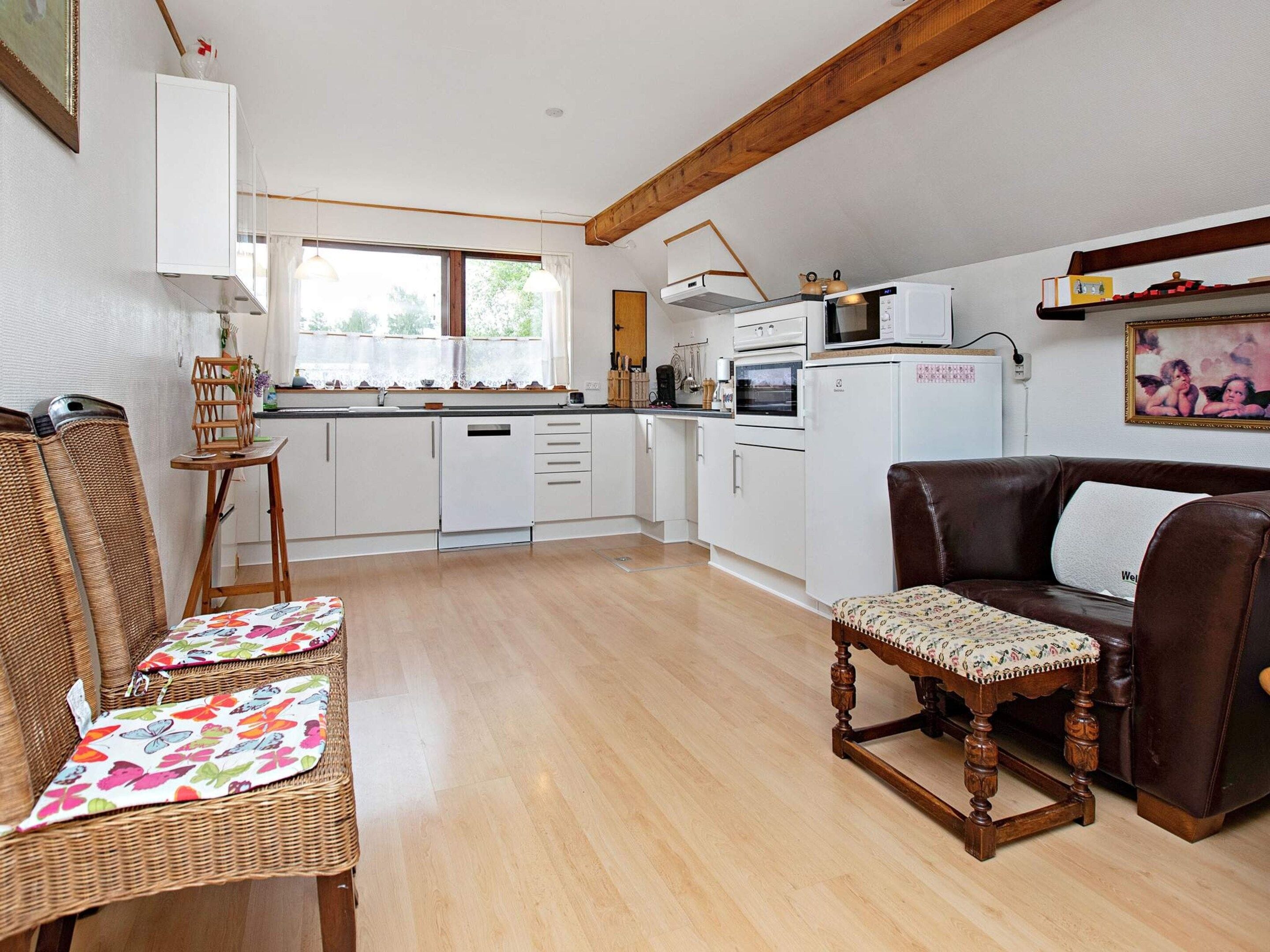 A compact kitchen with white cabinets, a microwave, and a refrigerator. There is a wooden bench with a cushion and a brown leather sofa with a cushion.