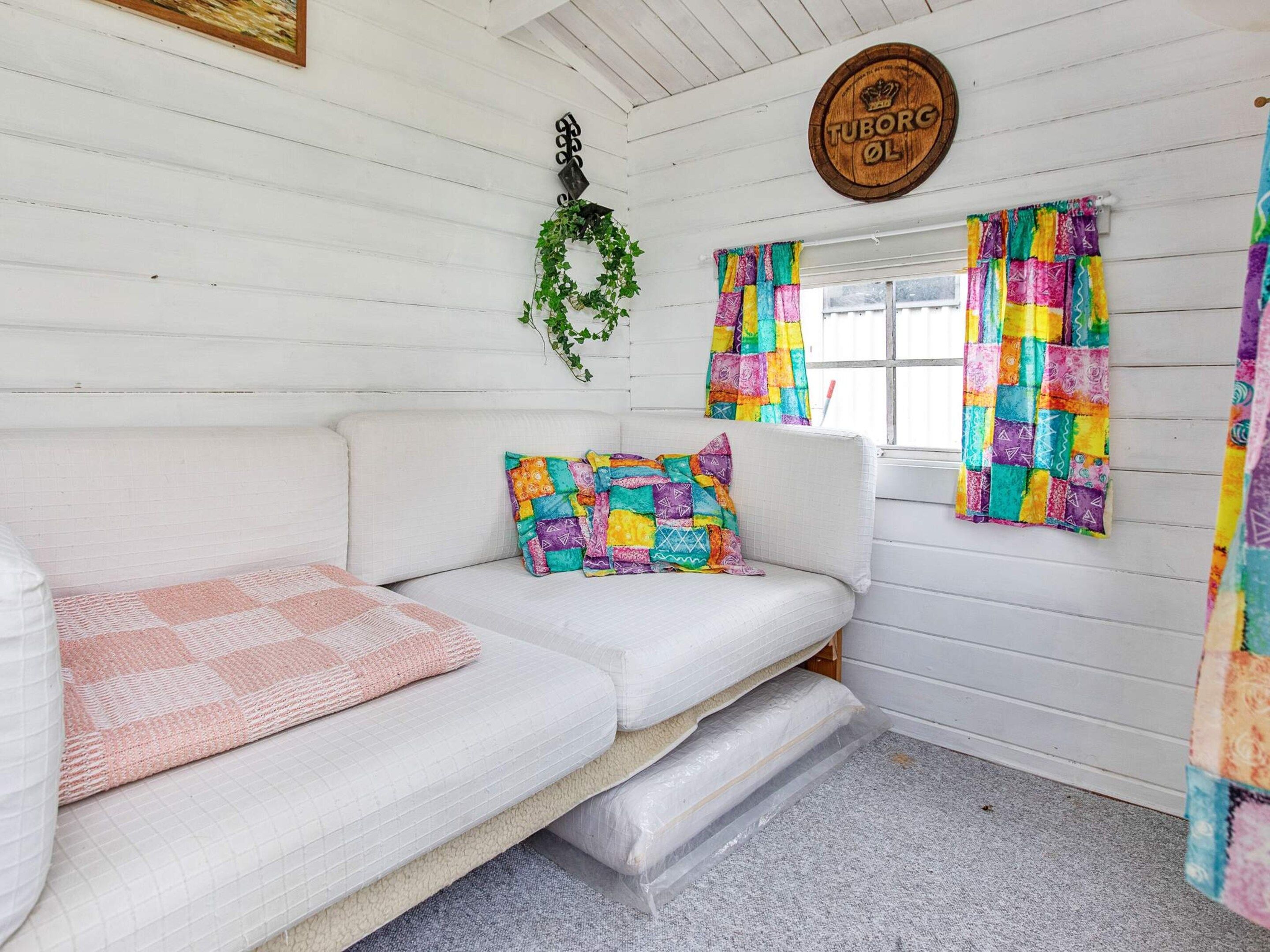 A cozy room with a white sofa, colorful pillows, a hanging plant, and a window with colorful curtains.