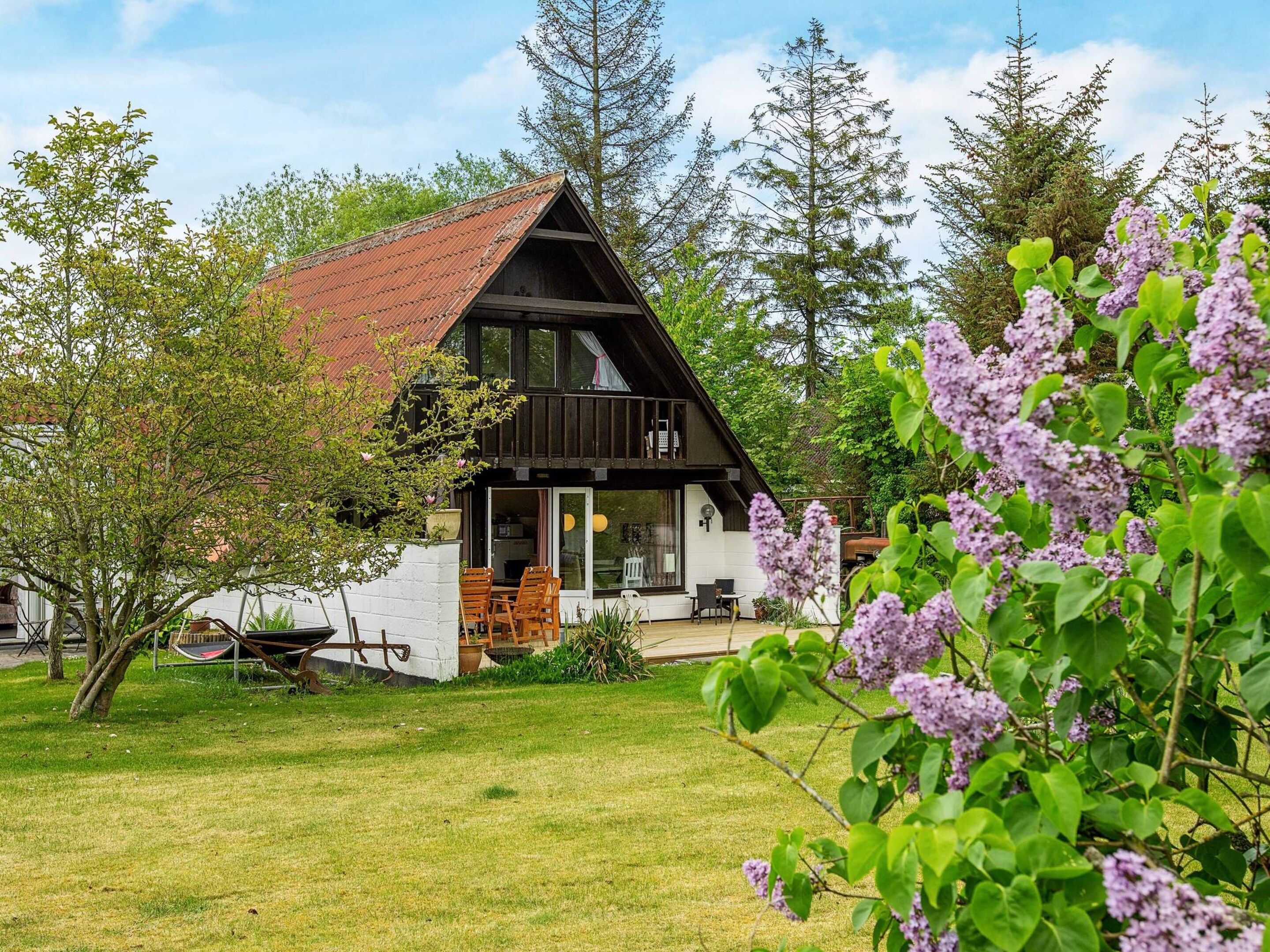 En stuga med ett sluttande tak, en veranda och en trädgård med träd och blommor.