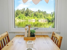 Mesa de madeira com cadeiras, janela com vista para um lago e árvores, e objetos decorativos no parapeito.