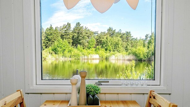 A wooden dining table with chairs, a window with a view of a lake and forest, and decorative items on the windowsill. - 4 Person Holiday Home in Laeso (Læsø)