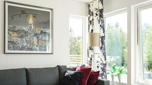 Un salon moderne avec un canapé gris, une table basse en verre, un tableau représentant un paysage urbain encadré et une fenêtre ornée de rideaux à motifs floraux.