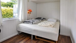 A small, single-bed room with a wooden floor, white walls, a window with white curtains, and a wall-mounted lamp. - Holiday Home in Fjällbacka (Faxe Ladeplads)