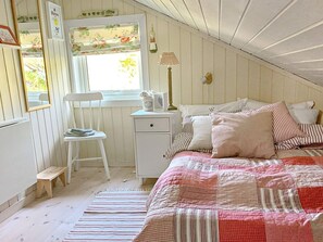 Um quarto de sótão aconchegante, com teto inclinado, uma escrivaninha de madeira com cadeira, uma cama com roupa de cama listrada e uma janela com persianas florais.