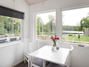 Una mesa de comedor blanca con dos sillas, un florero con flores y vistas a un lago y un bosque a través de ventanas amplias.
