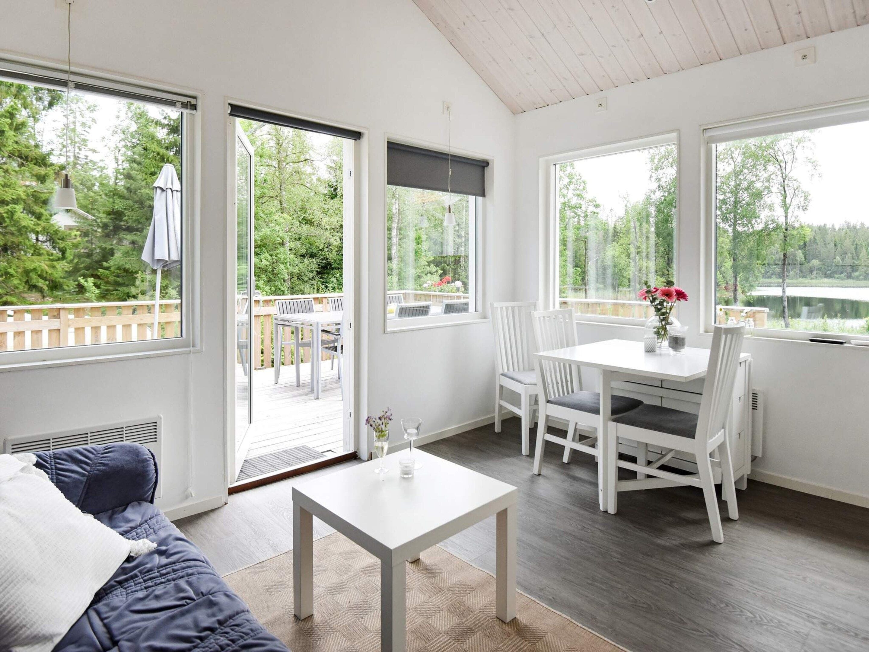 Quarto com mesa e cadeiras brancas, sofá e janelas amplas com vista para um deck e área verde.