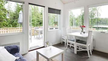 A room with a white table and chairs, a sofa, and large windows offering a view of a deck and greenery.