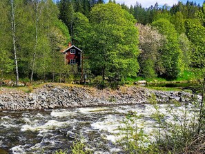 Una casa junto a un río con una ribera rocosa y un bosque denso al fondo.