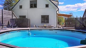 A swimming pool with a wooden deck, a covered patio, and a white building with windows.