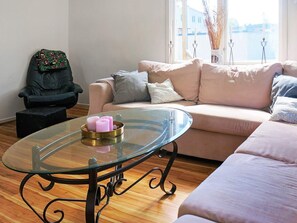 A living room with a glass-top coffee table, a sofa, a black leather chair, and a window with a view of a building and a body of water. - 7 Person Holiday Home in Billingsfors (Billingsfors)