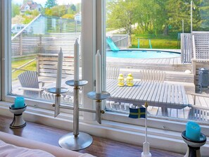 Visão pela janela de uma área de piscina com deck, móveis de exterior e uma bandeira.