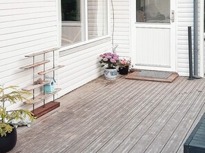 Une terrasse en bois avec une porte blanche, un petit espace porche et un pot de fleurs contenant une plante aux fleurs roses.