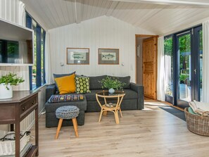 A cozy living room with a grey sofa, a small round table, and a wicker chair. There are two framed fish pictures on the wall, a potted plant, and a wooden door leading outside. - 5 Person Holiday Home in Grenaa (Grenaa)