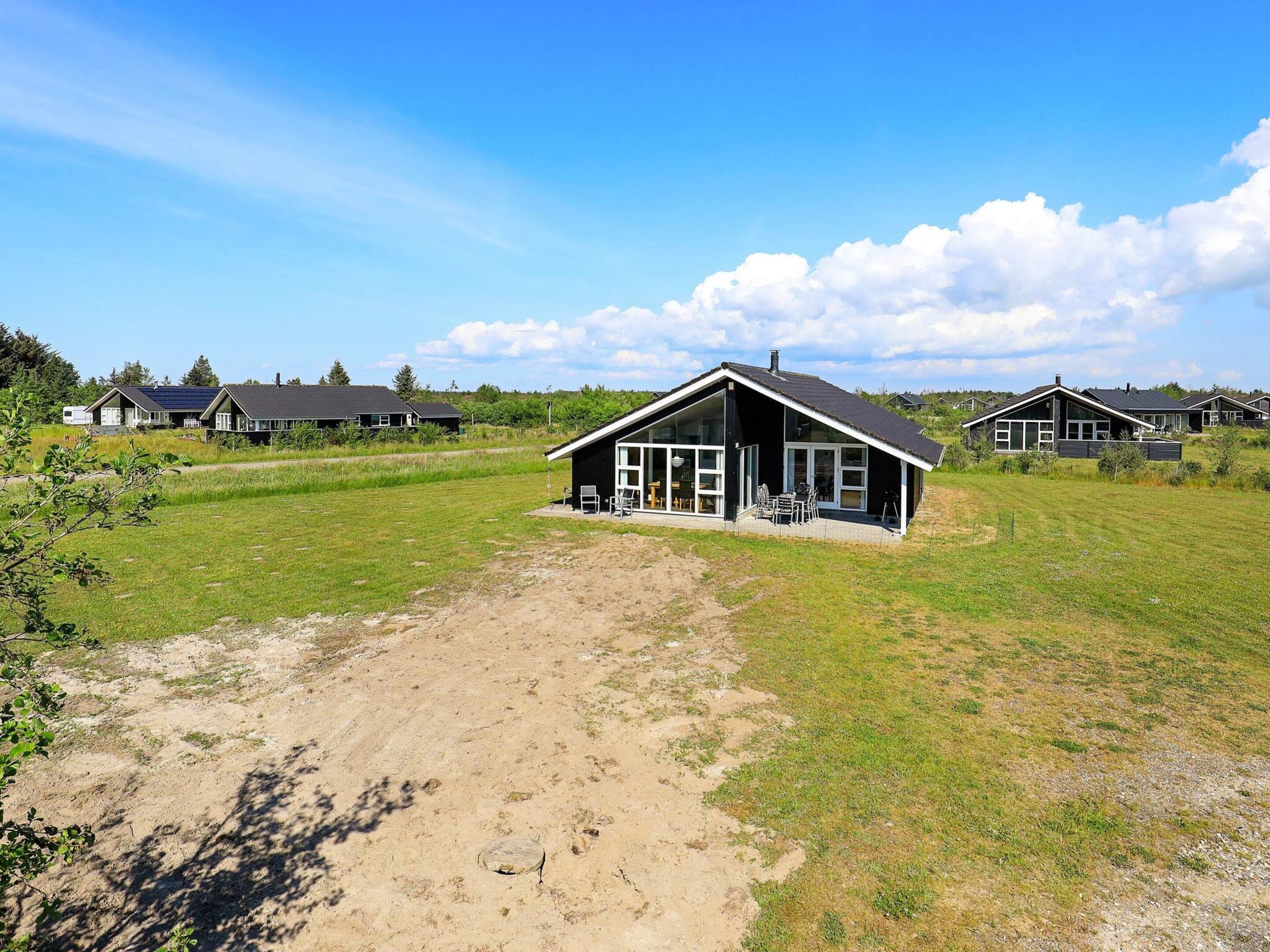 A modern house with a large patio area, surrounded by a grassy field and other similar houses in the background.