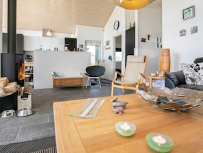 A modern living room with a wooden coffee table, a black sofa, a wicker chair, and a dining table with a vase and candles. - Holiday Home in Brovst (Brovst)