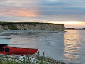 A calm river with a red boat docked on the shore, a grassy cliff, and a sunset in the background.