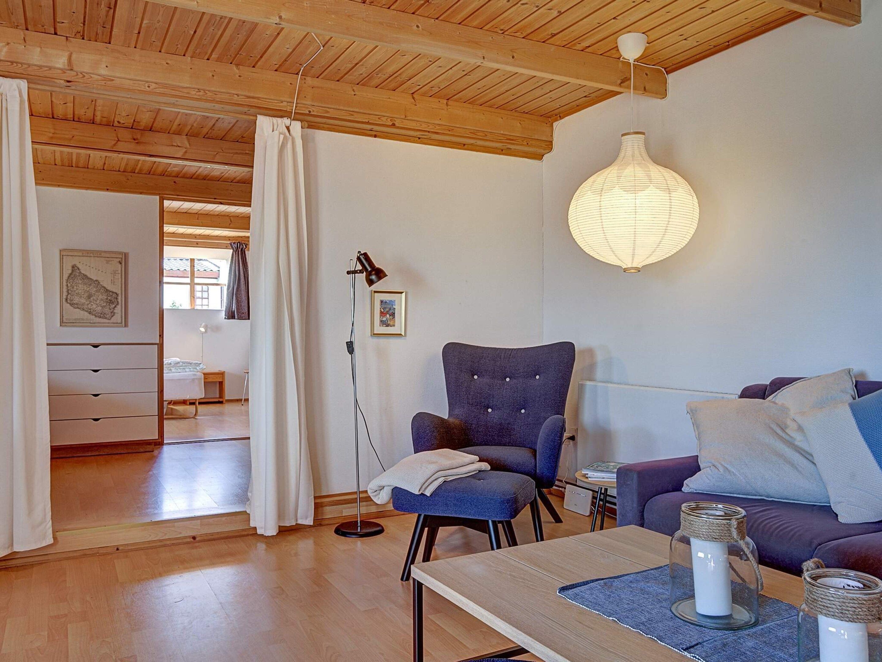 A cozy living room with a wooden ceiling, a blue armchair, a sofa, a table with candles, and a floor lamp.