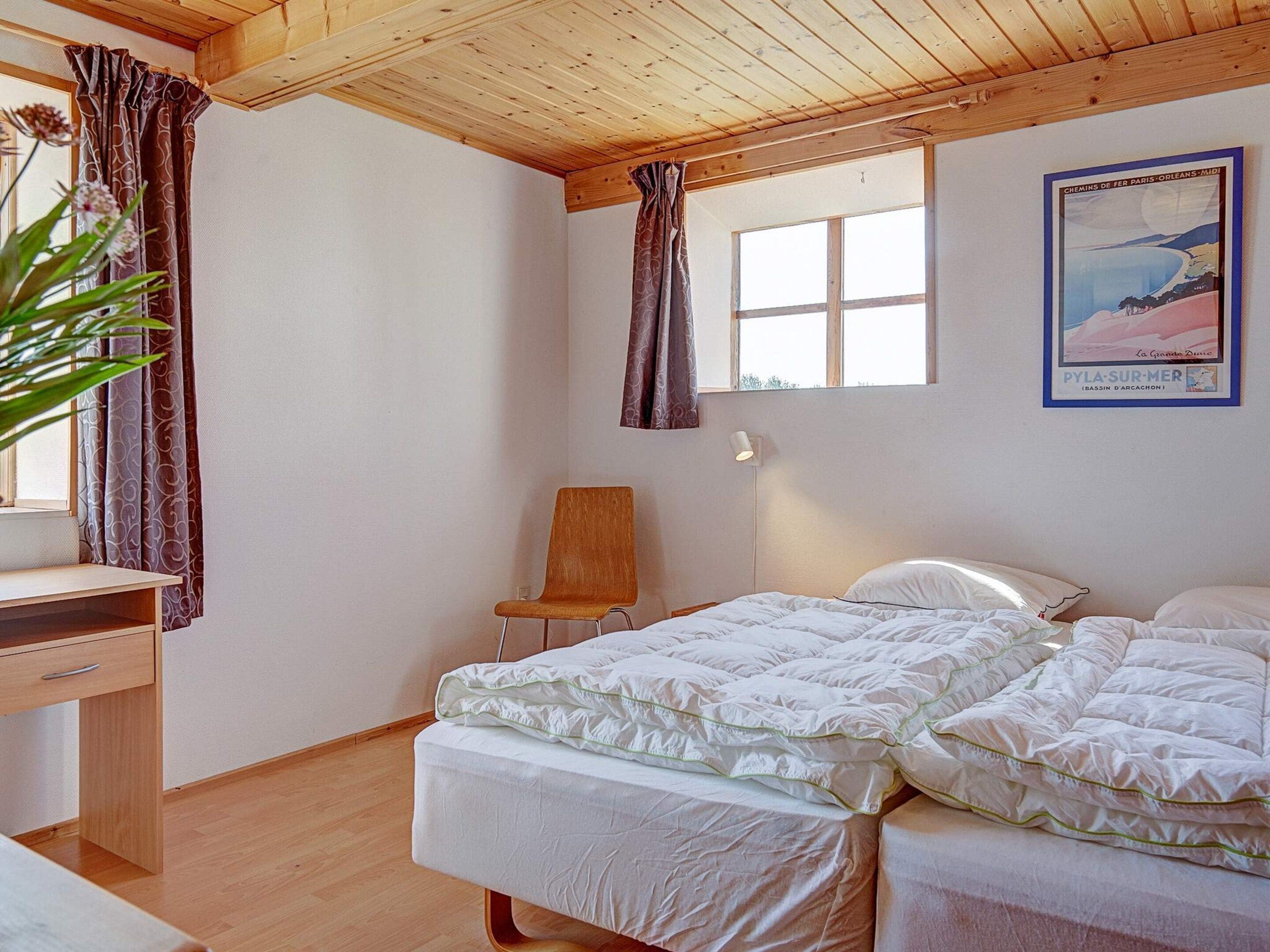 A room with a wooden ceiling, a bed with white bedding, a desk with a chair, and a window with curtains.
