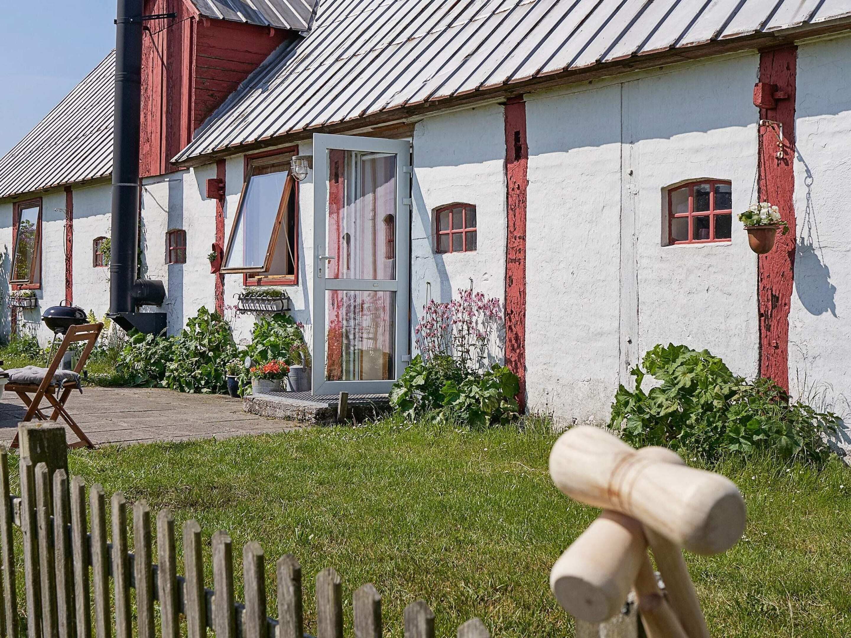 A rustic building with a white exterior and red accents, a wooden fence, and a garden area with potted plants.
