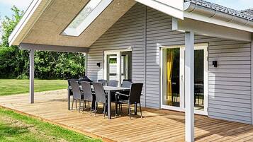 Un patio techado con una mesa y sillas, una terraza de madera y una casa con fachada gris y detalles blancos.