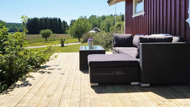 A wooden deck with outdoor furniture, including a sofa and ottoman, overlooking a scenic landscape with trees and a distant building. - 4 Person Holiday Home in Ucklum (Ucklum)