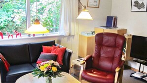 A living room with a black sofa, a red leather chair, a wooden coffee table with a bowl of fruit, and a window with a view of greenery.