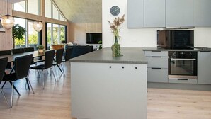 A modern kitchen with grey cabinets, a central island, and a dining area with a large window. - Holiday Home in Juelsminde (Juelsminde)