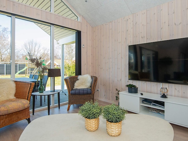 A modern living room with a large TV, wicker furniture, and a view of a playground. - Holiday Home in Juelsminde (Juelsminde)
