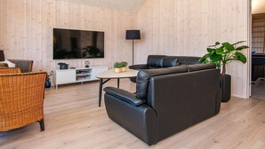 A modern living room with a large flat-screen TV, a black leather sofa, a wicker chair, and a small table with plants. - Holiday Home in Juelsminde (Juelsminde)