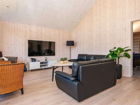 A modern living room with a large flat-screen TV, a black leather sofa, a wicker chair, and a small table with plants. - Holiday Home in Juelsminde (Juelsminde)