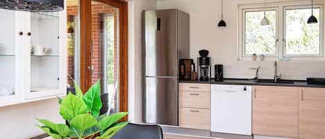 A modern kitchen with a stainless steel refrigerator, a coffee maker, and a dishwasher. There is a dining table with a potted plant in the foreground.
