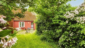 Ein rotes Haus mit Ziegeldach, ein kleiner Garten mit Rasen und einem Tisch mit Stühlen, sowie ein dichter Heckenstreifen mit rosa Blüten.
