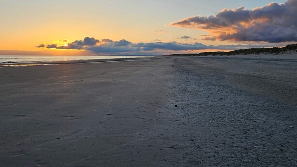 Ein Strand mit Sonnenuntergang, sandigem Ufer und Dünen im Hintergrund.