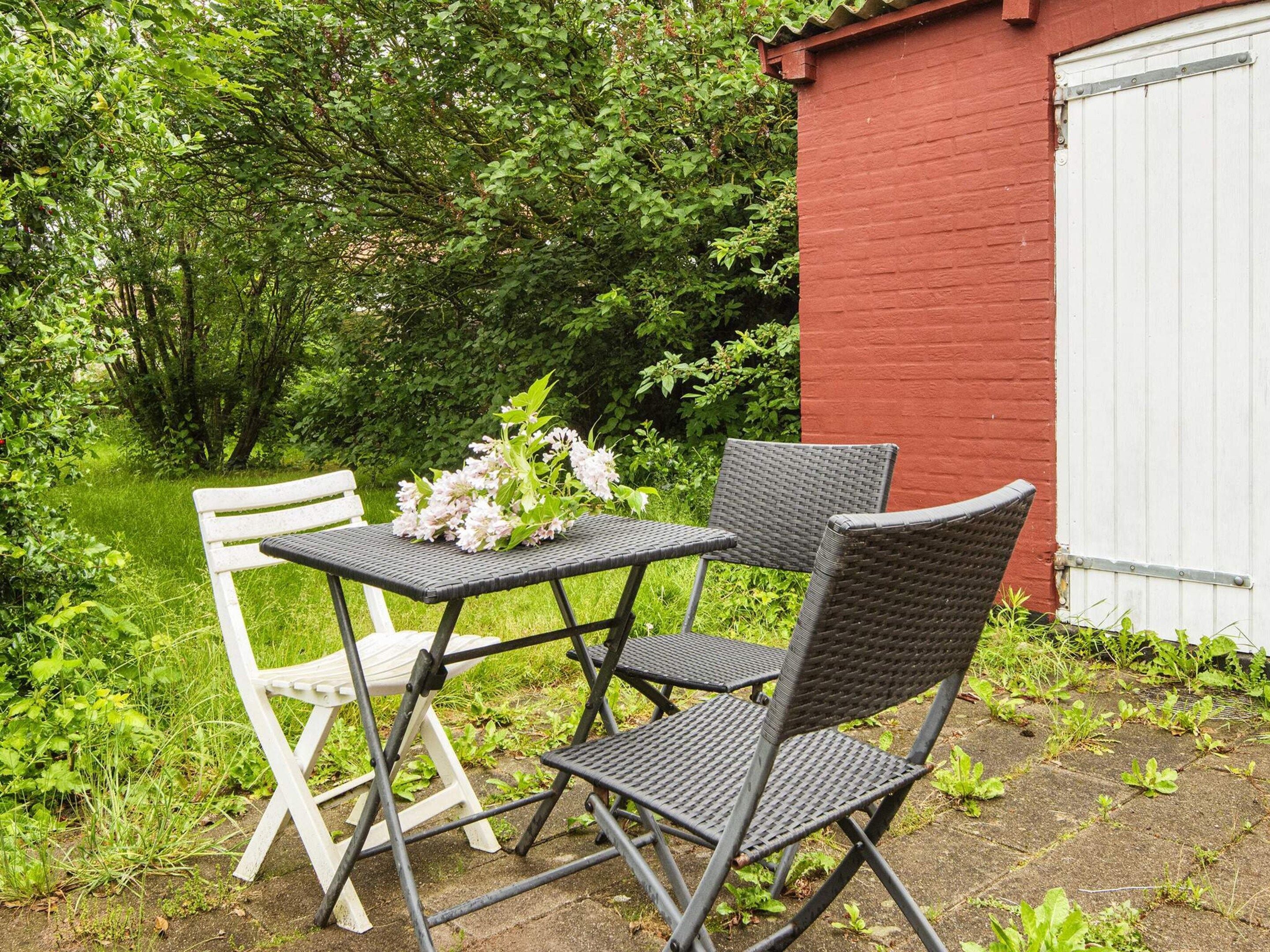 An outdoor seating area with a table and chairs, a small garden, and a red building in the background.