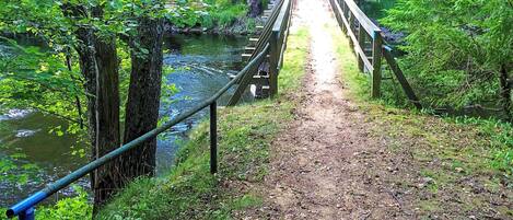 A wooden bridge over a river with a dirt path leading to it, surrounded by trees and greenery.