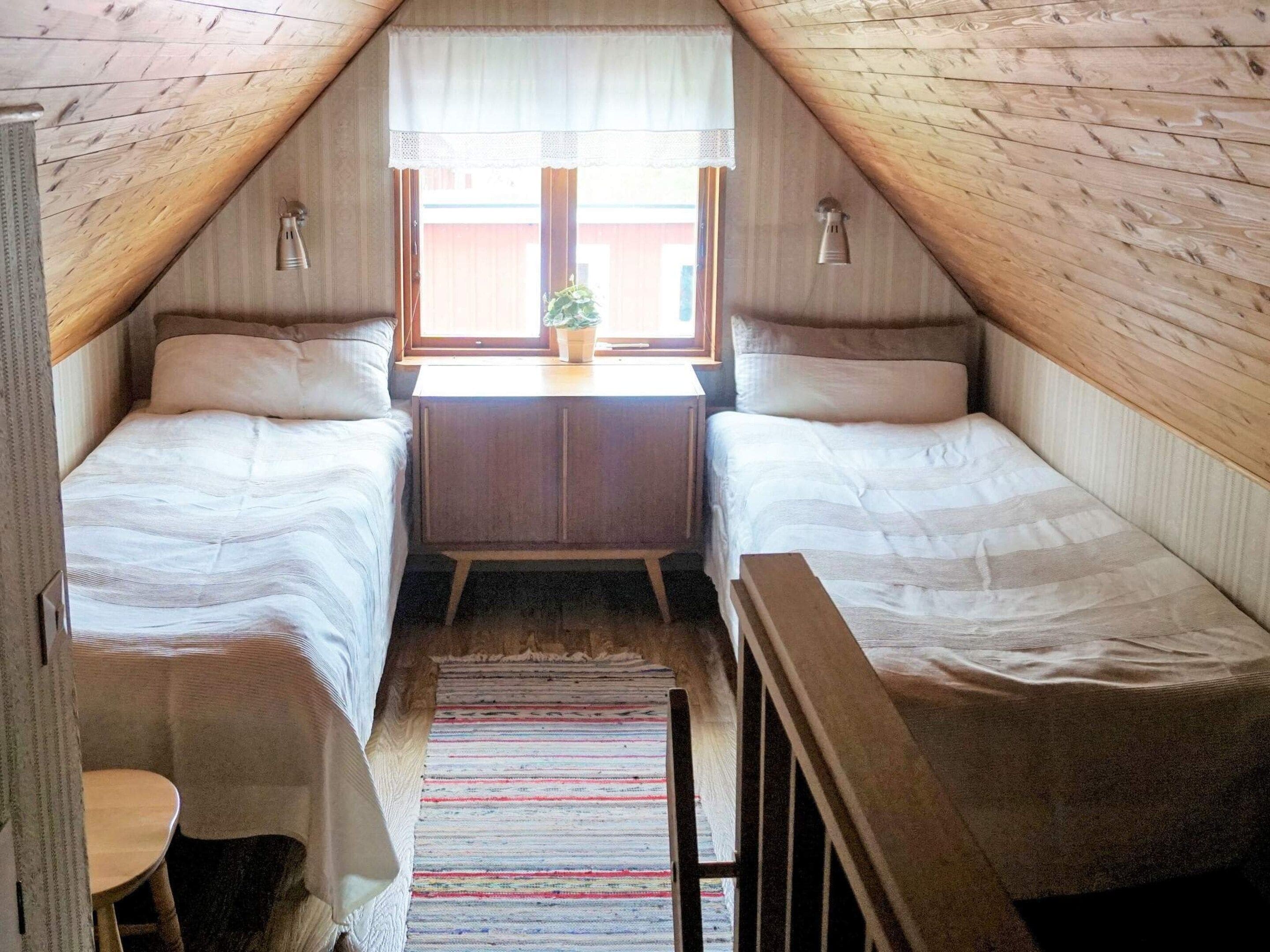 Quarto de dormitório com duas camas individuais, uma mesa pequena, uma cadeira, uma janela com planta e teto de madeira.
