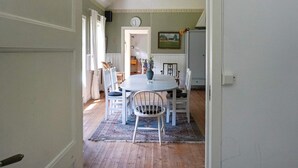 A dining area with a round table, white chairs, and a rug. There is a clock on the wall and a painting above the door.