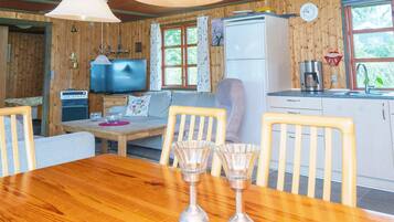 Interior acolhedor de uma cabana com uma mesa de jantar de madeira, cadeiras, um sofá, uma televisão, um frigorífico e uma cozinha com pia e fogão.