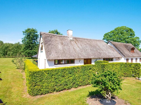 A white house with a thatched roof, surrounded by a well-maintained hedge and a grassy lawn.