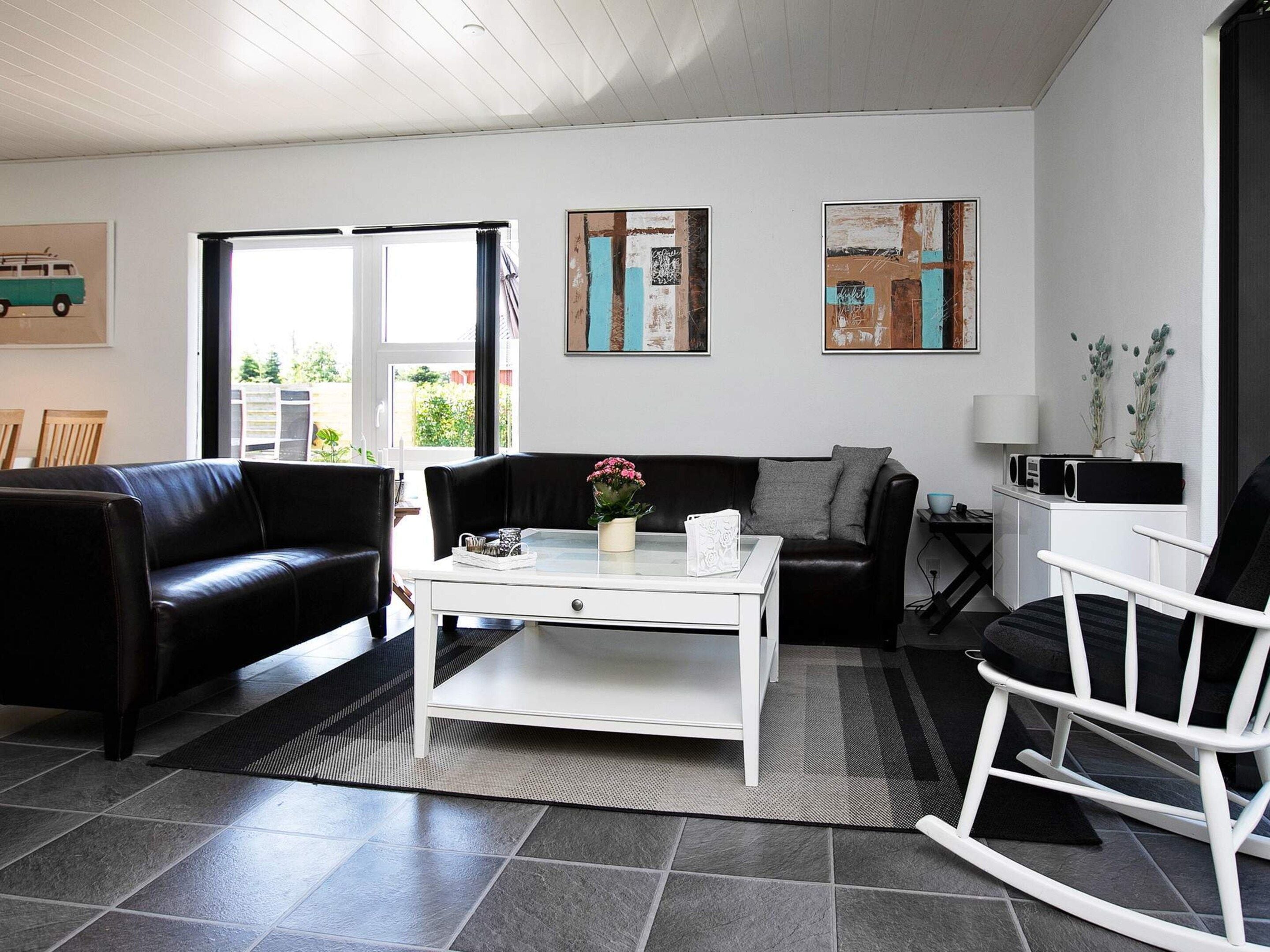 Un salón con un sofá de cuero negro, una mesa de centro blanca y un sillón mecedor blanco. Hay dos cuadros abstractos en la pared y una ventana con vistas a zonas verdes.