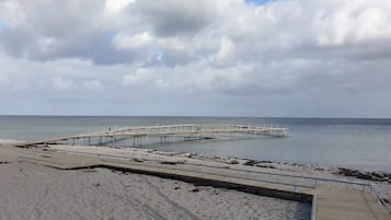 Una playa con un muelle de madera que se adentra en aguas tranquilas, rodeada de arena y bajo un cielo nublado.
