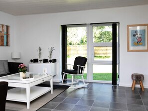 A modern living room with a white sofa, a dining table, a black rocking chair, and a small stool. There is a window with a view of a garden.