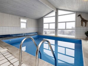 An indoor swimming pool with clear blue water, metal ladders, and large windows offering views of the outside.