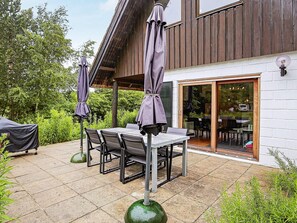 An outdoor patio with a table and chairs, surrounded by greenery, and a building with a wooden facade and glass doors.