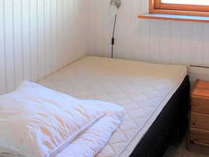 Una cama individual con colchón y almohada blancos, una mesita de noche con cajones y una ventana con persianas.