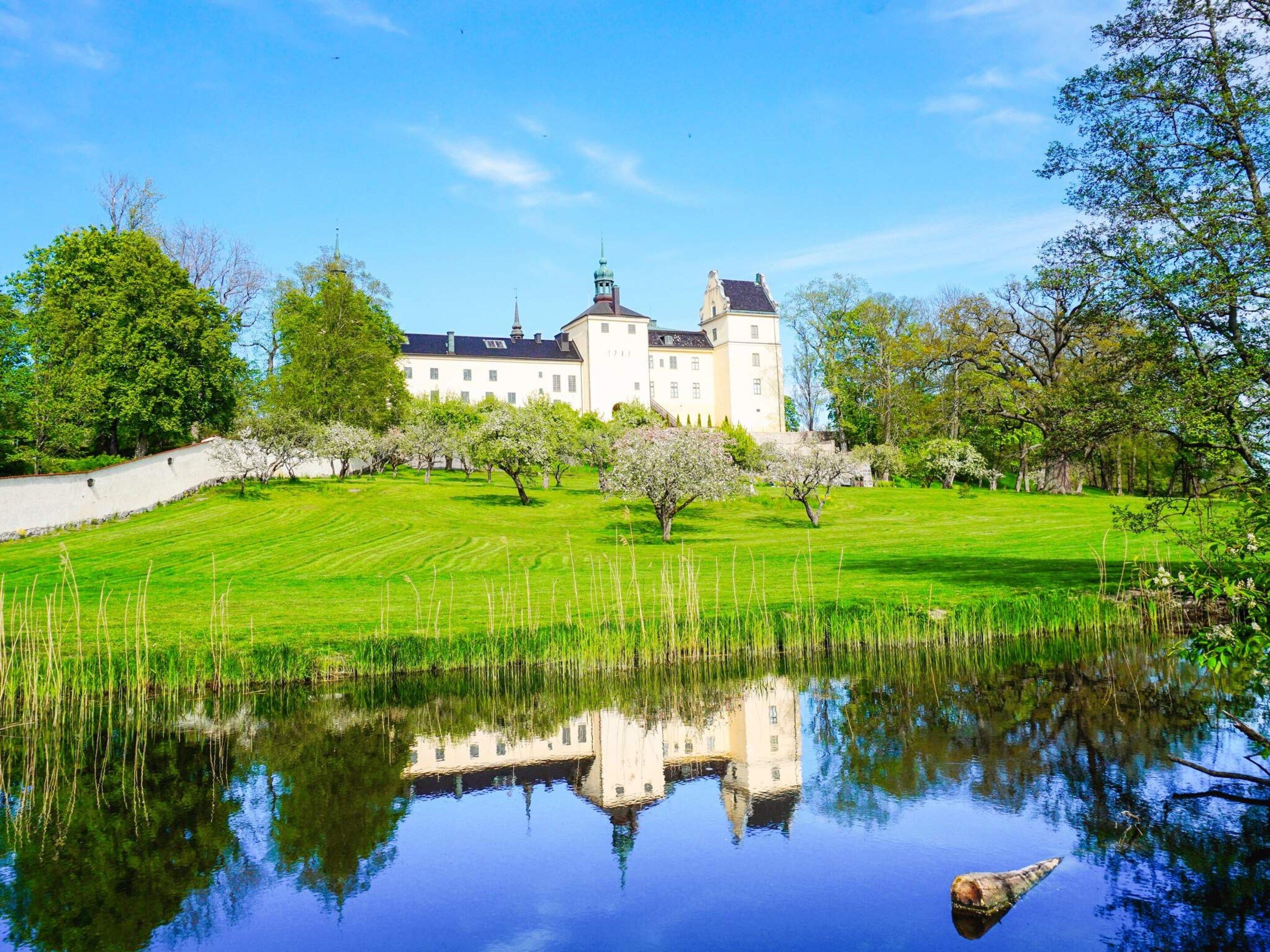 Un grand bâtiment blanc à la façade symétrique, entouré d’une pelouse bien entretenue et bordé d’une étendue d’eau dans laquelle se reflète le bâtiment.