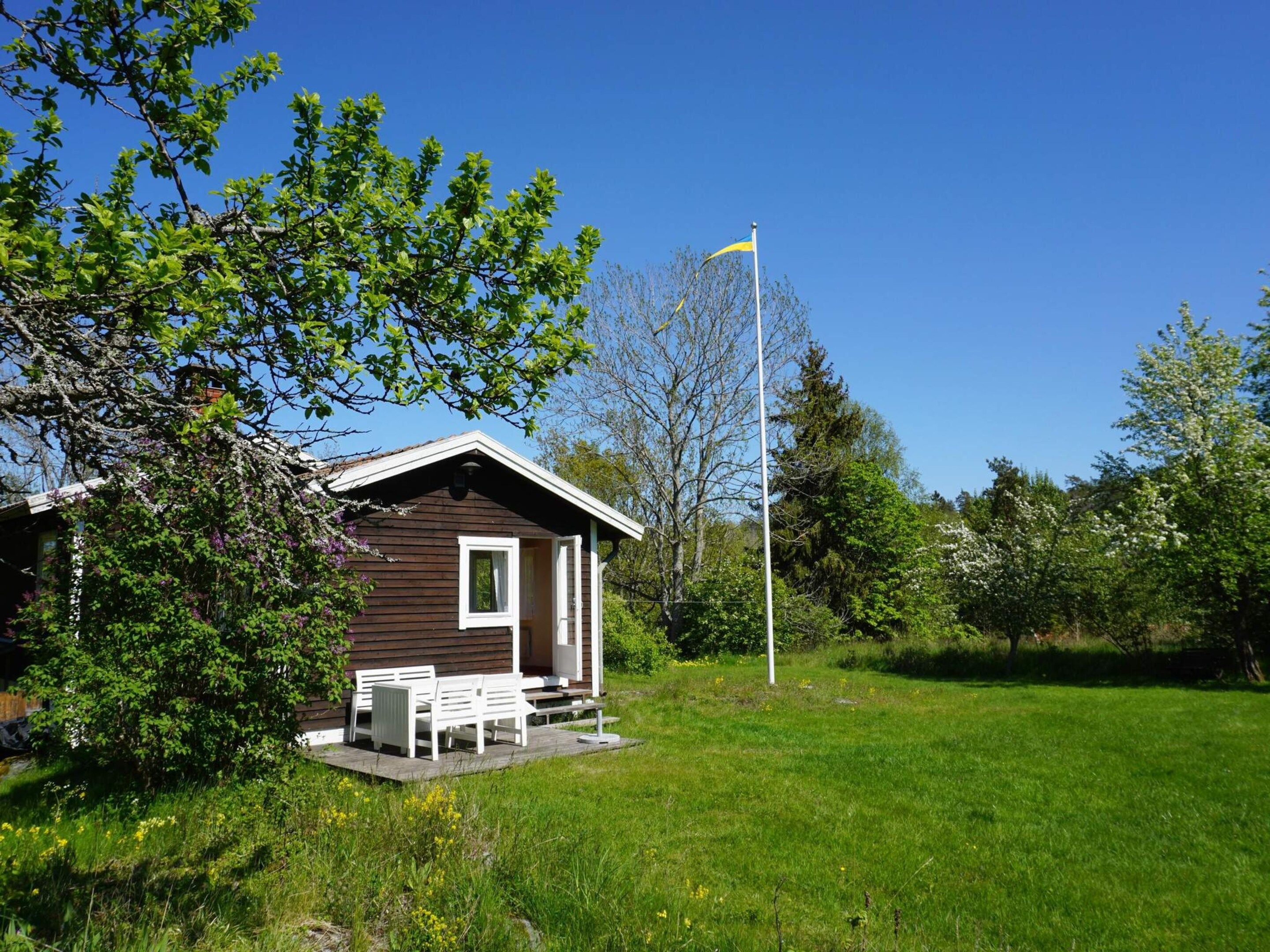 Eine kleine Holzhütte mit Veranda und Sitzgelegenheit, umgeben von üppiger Vegetation und blauem Himmel.