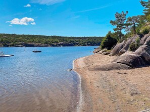 A clear view of a beach with a rocky shoreline, calm water, and a forested background. - 9 Person Holiday Home in Vesteroy (Hvaler)