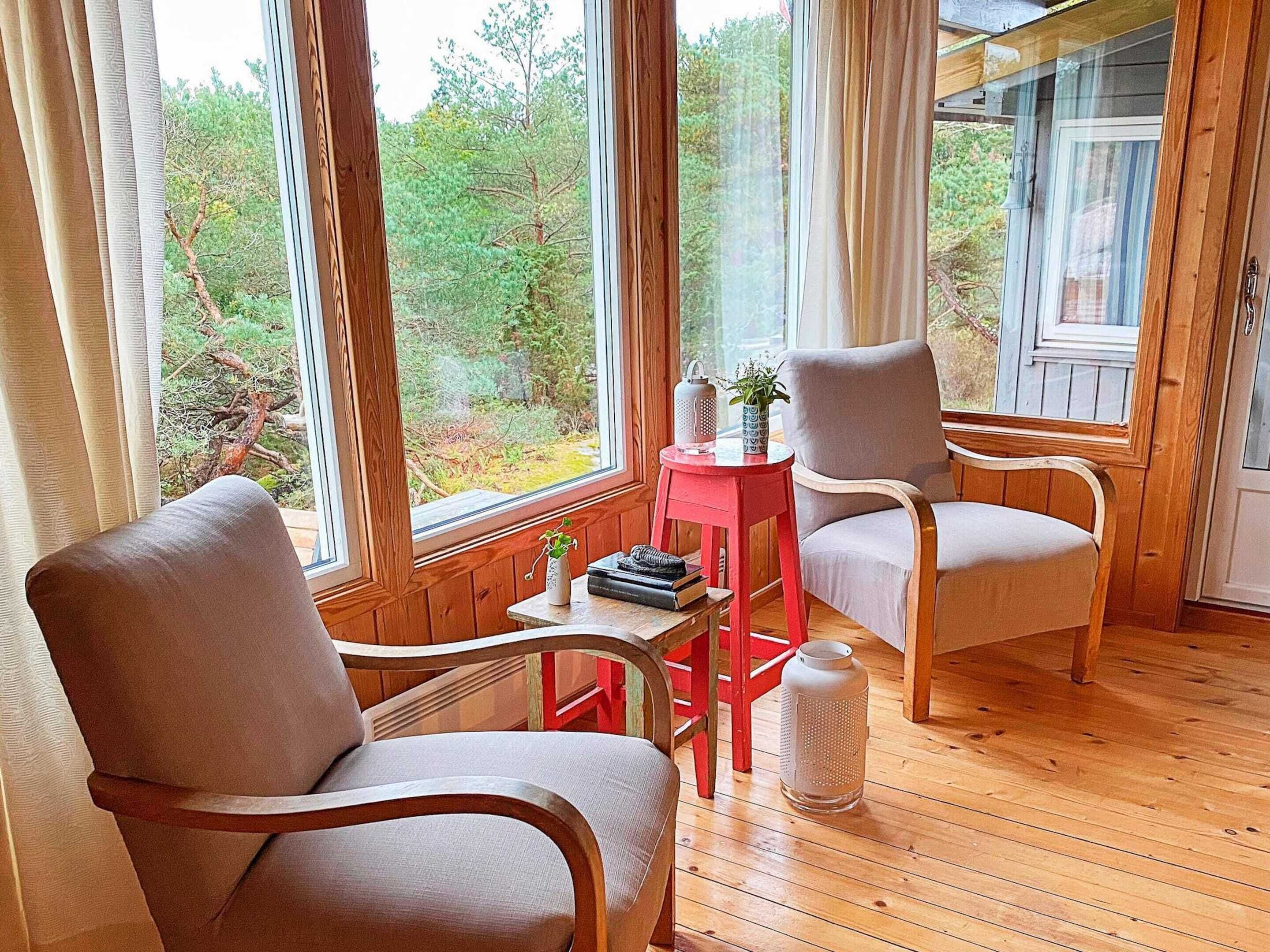 Une pièce chaleureuse avec un plancher en bois, deux fauteuils, une petite table rouge ornée d’une plante, et une vue sur une forêt à travers de grandes fenêtres.