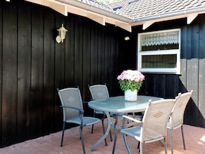 An outdoor patio with a round table and four chairs, a potted plant with flowers, a wall-mounted lantern, and a window with lace curtains. - Holiday Home in Hadsund (Hadsund)
